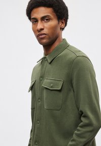 Camicia a maniche lunghe verde con tessuto testurizzato, due tasche sul petto e colletto, con chiusura frontale a bottoni.