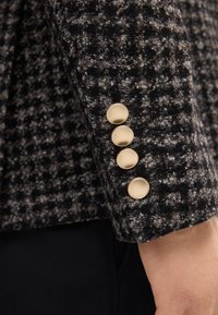 Czarna i szara tkanina w wzór houndstooth o teksturowanym wykończeniu. Cztery złote guziki na mankiecie. Elegancki, dopasowany krój.