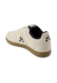 Scarpa sportiva beige chiaro con una texture in suede liscia, accenti neri e suola in gomma color gomma. Presenta un logo sul lato e dettagli perforati.