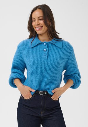 Femme souriante, portant un pull bleu en tricot à trois boutons et un jean foncé, les mains dans les poches, debout devant un fond blanc uni.