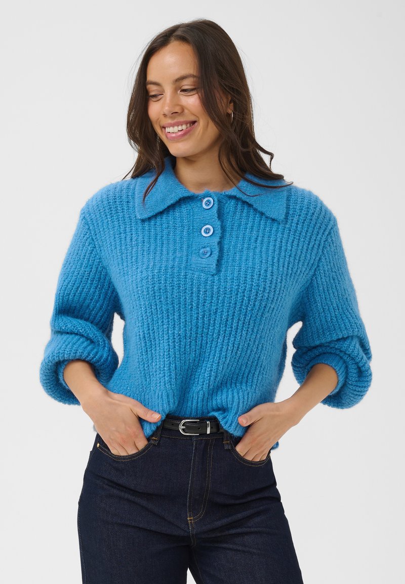 Femme souriante, portant un pull bleu en tricot à trois boutons et un jean foncé, les mains dans les poches, debout devant un fond blanc uni.