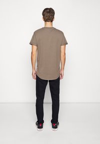 Homme se tenant devant un mur blanc, portant un t-shirt court brun, un pantalon noir et des baskets noires avec des semelles blanches et des détails rouges.