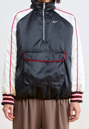 Veste de sport Nike noire et blanche avec bordure rouge, col zippé, poche avant et poignets rayés portée avec un pantalon marron.
