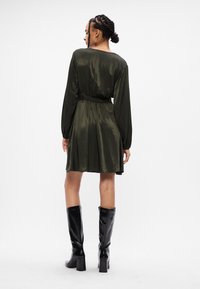 Ελιά πράσινο φόρεμα με μακριά μανίκια, σφικτή μέση και ρευστή φούστα. Διαθέτει λείο, γυαλιστερό ύφασμα και συνδυάζεται με μαύρες μπότες ως το γόνατο.