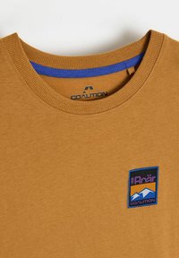 Koszulka z krótkim rękawem w musztardowym kolorze z niebieskim ściągaczem na dekolcie i haftowaną, wielokolorową łatką z logo na piersi. Miękka tkanina, casualowy krój.