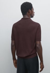 Kortærmet brun polo-shirt med ribbet tekstur, sideslidser og en strikket krave. Båret med sorte bukser på en neutral baggrund.
