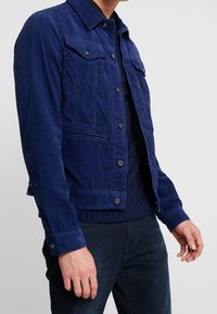 Man i blå manchesterjacka med knäppta fickor över en mörkblå ribbad tröja och mörkblå byxor.