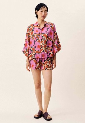 Vrouw staand, gekleed in een loszittend, kleurrijk bloemenoverhemd met knoopsluiting en bijpassende korte broek, met donkere open-toe slippers, tegen een effen achtergrond.