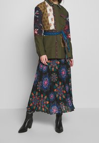 Olijfgroene patchwork blouse met bloemenaccenten, omgeven door een blauwe gestructureerde riem, gecombineerd met een zwarte plissérok met kleurrijke bloemenontwerpen.