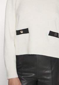 Pull tricot blanc avec poches décoratives noires et boutons dorés, porté avec un pantalon en cuir noir, montrant le torse et le bras gauche.