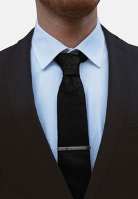 Schwarte Krawatte mit einem Rautenmuster, silberner Krawattenclip, getragen über einem hellblauen Hemd und einer dunklen Anzugjacke. Strukturierte Stoffdetails sichtbar.