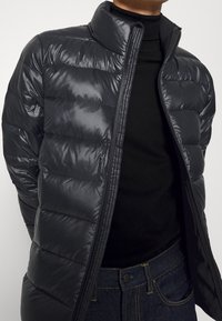 Persona vistiendo una chaqueta acolchada negra brillante sobre un suéter de cuello alto negro y jeans azul oscuro, con la chaqueta parcialmente desabrochada.