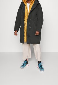 Parka noire avec capuche et cordon de serrage ajustable, portée sur un gilet matelassé jaune. Pantalon de couleur claire et baskets bleues avec des accents roses.