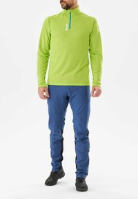 Chemise longue à manches longues, texturée et vert vif avec un col zippé. Associée à un pantalon bleu présentant des accents latéraux noirs et des chaussures noires robustes.
