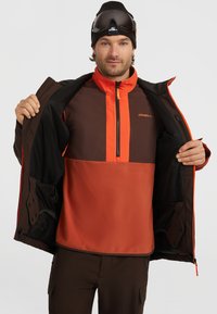 Bruine en oranje skijas met een ritssluitingen, contrasterende kleurvlakken en een merkl logo. Model draagt een skibril en een beanie.
