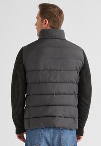 Gepolsterte Weste aus dunkelgrauem Material mit hohem Kragen. Verfügt über einen strukturierten Strickpullover an den Ärmeln und ein horizontales Steppdesign.