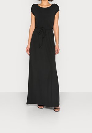 Kobieta w czarnej sukience maxi bez rękawów z przewiązanym paskiem w talii i czarnych sandałach z odkrytymi palcami, stojąca na jednolitym tle.