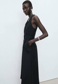 Black sleeveless dress with a v-neckline, waist belt, side pockets, and a flowing silhouette. Smooth fabric and minimalist design.