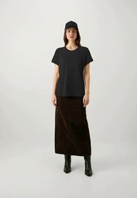 Kvinne som har på seg svart caps, svart t-skjorte med korte ermer, mørkebrun lang skjørt og svarte ankelstøvler, stående mot en enkel lys bakgrunn.