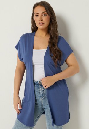Donna che indossa un cardigan blu aperto su una maglietta bianca e jeans azzurri, in piedi contro un muro semplice.