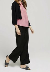 Schwarzer, kurzer Cardigan über einer rosa, gemusterten Bluse mit Knöpfen, kombiniert mit schwarzen, weit geschnittenen Hosen und dunklen Sandalen zum Hineinschlüpfen.