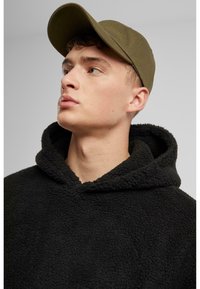 Haut à capuche en polaire noire avec une surface texturée, associé à une casquette vert olive. La casquette présente un bord courbé et un design simple.