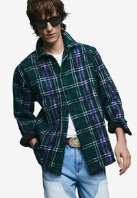 Grün-schwarzes kariertes Hemd mit lila und weißen Akzenten, Knopfleiste vorne, kragenloses Design, kombiniert mit hellblauen Jeanshosen.