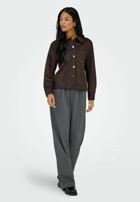 Camisa de ante marrón con botones y dos bolsillos frontales, combinada con pantalones anchos grises y zapatos negros. El modelo posa en un fondo neutro.
