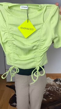 Svijetlozeleni rebrasti top s kratkim rukavima, s nabrusima sprijeda i vezicama, koji nosi osoba u bež hlačama s prugicama.