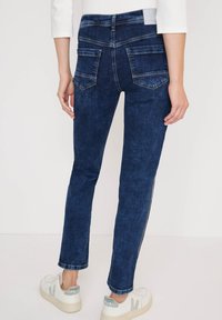 Jeans de mezclilla azul oscuro con corte ajustado, con bolsillos traseros y rayas blancas laterales. Combinados con zapatillas blancas.