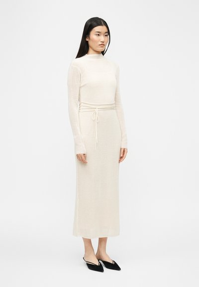 Femme aux longs cheveux foncés portant une robe midi en maille côtelée crème à manches longues avec ceinture à la taille et chaussures plates noires à bout pointu.