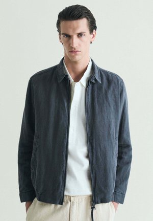 Joven con el cabello oscuro peinado hacia atrás, vistiendo una camisa polo blanca bajo una chaqueta gris oscuro y pantalones beige, de pie contra un fondo liso.