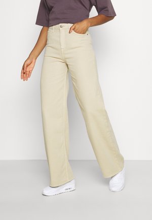 Persona con pantalones anchos beige, zapatillas blancas y una camiseta de manga corta de color púrpura oscuro, con una mano en el bolsillo y la otra a un lado.