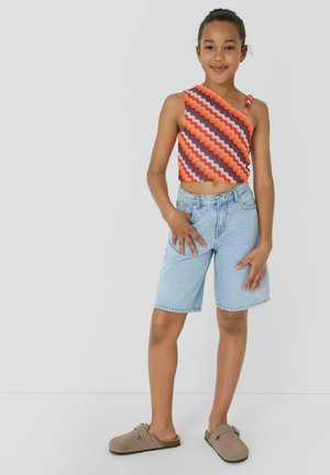 Dziewczyna stoi, ubrana w asymetryczny top z wzorem zygzaków, jasne krótkie spodenki jeansowe oraz taupe mokasyny na białym, jednolitym tle.