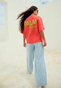 Korálová oversized tričko s modrým potiskem "PALM Springs" na zádech, spárované s světle modrými pruhovanými širokými kalhotami.