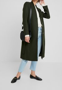 Cappotto lungo verde scuro con una vestibilità rilassata, tessuto morbido e bottoni frontali, abbinato a jeans azzurri chiari e mocassini neri. Piccola borsa a tracolla nera.