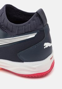 Scarpa sportiva navy con colletto in maglia, tomaia in mesh e materiale sintetico, dettagli con strisce bianche e suola in gomma rossa. Logo Puma visibile.