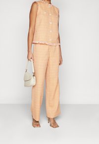 Meruňkový tweedový dvoudílný oblek s třaslavými okraji, obsahující top bez rukávů a široké kalhoty, doplněný lehkou kabelkou a zlatými sandály.