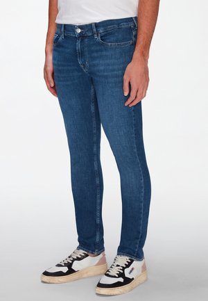 Hombre con vaqueros ajustados azules y zapatillas blancas con detalles en negro y rosa, de pie contra un fondo blanco liso.