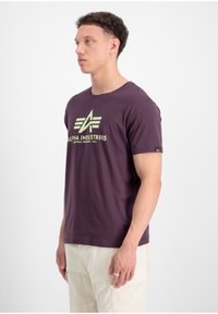 Lila Kurzarm-T-Shirt mit einem großen grünen Logo mit der Aufschrift "ALPHA INDUSTRIES." Aus Baumwollstoff mit lässigem Schnitt. Einfaches Design.
