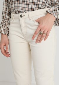 Béžové džíny s vysokým pasem a slim fit, vyrobené z denimu. Mají přední zapínání na knoflík a postranní kapsy. Detailní záběr na ruku, která nosí prsteny.