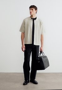 Νεαρός άνδρας που φοράει μπεζ πουκάμισο με κοντά μανίκια πάνω από μαύρο μπλουζάκι και παντελόνι, κρατώντας μια μαύρη δερμάτινη τσάντα, στέκεται σε εσωτερικό χώρο.