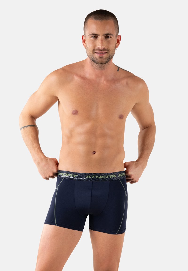 Homme torse nu ajustant un caleçon bleu marine avec la ceinture "ATHETA SPORT", debout devant un fond blanc uni.