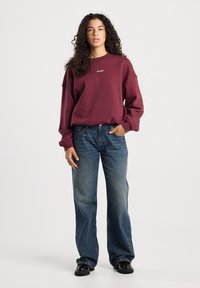 Vrouw met krullend haar, gekleed in een bordeauxrode sweatshirt met de tekst "New York", blauwe wijdpijpen jeans en zwarte schoenen, staand tegen een effen achtergrond.