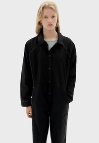 Czarna zapinana na guziki kurtka z kołnierzem, z kontrastującym białym przeszyciem, luźnym krojem i dwoma przednimi kieszeniami. Gładka faktura.
