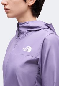 Άτομο που φοράει ένα ανοιχτό μωβ μπουφάν North Face με φερμουάρ και ψηλό γιακά, με ορατό λογότυπο στο στήθος, εμφανιζόμενο από το πλάι και τους ώμους.