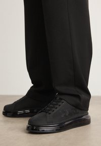 Juodi žemakulniai sportbačiai su storais skaidriais padais ir šone išgraviruotu "KARL", dėvimi su laisvais juodais kelnėmis ant betono grindų.