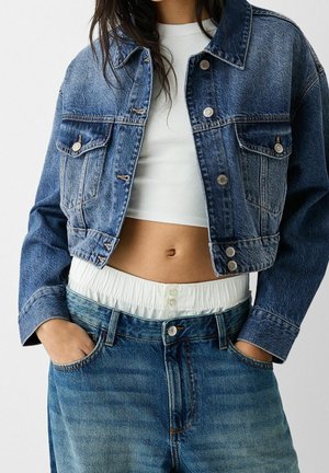 Woman wearing cropped two-tone denim jacket over white cropped top and high-waisted blue jeans with elastic waistband, hands in pockets.