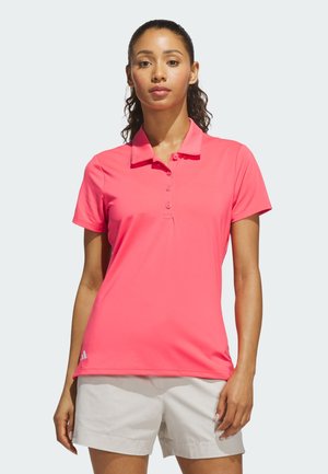 Femme portant un polo rose vif à manches courtes et un short beige clair, debout avec les bras détendus devant un fond uni.