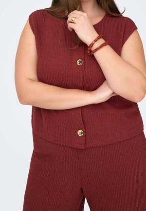 Personne portant un haut bordeaux côtelé sans manches et un pantalon assorti, avec des boutons dorés et des bracelets rouges en perles au poignet.